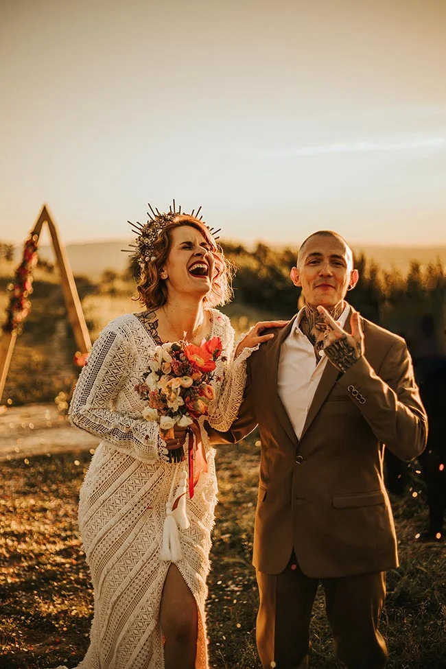 Couple riant lors d'un elopement au coucher du soleil, elle en robe bohème et couronne, lui en costume beige.