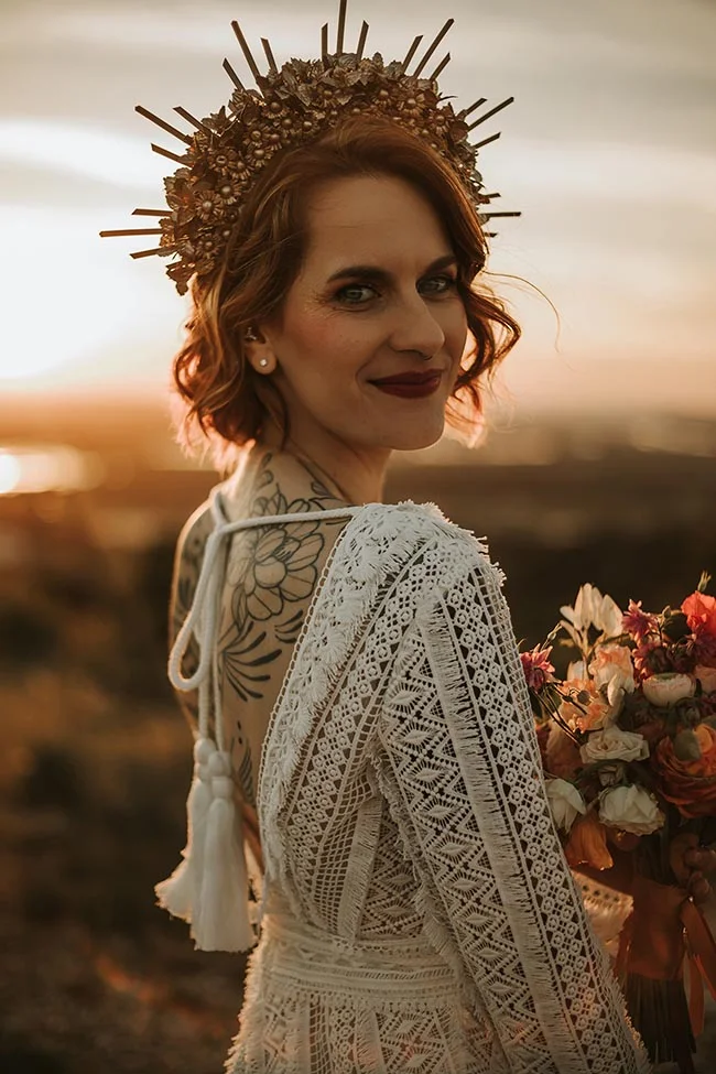 Mariée aux cheveux roux portant une couronne dorée et une robe en dentelle blanche, tenant un bouquet au coucher du soleil.