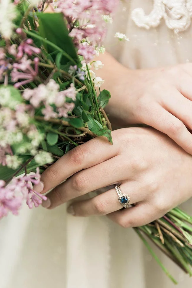 Gros plan sur les mains d'une mariée tenant un bouquet de fleurs roses et blanches, avec une bague en saphir bleu.