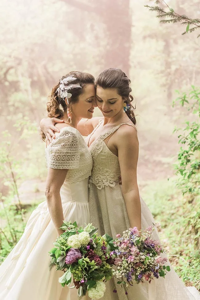 Deux mariées s'enlacent tendrement en forêt, vêtues de robes blanches et tenant des bouquets champêtres colorés.