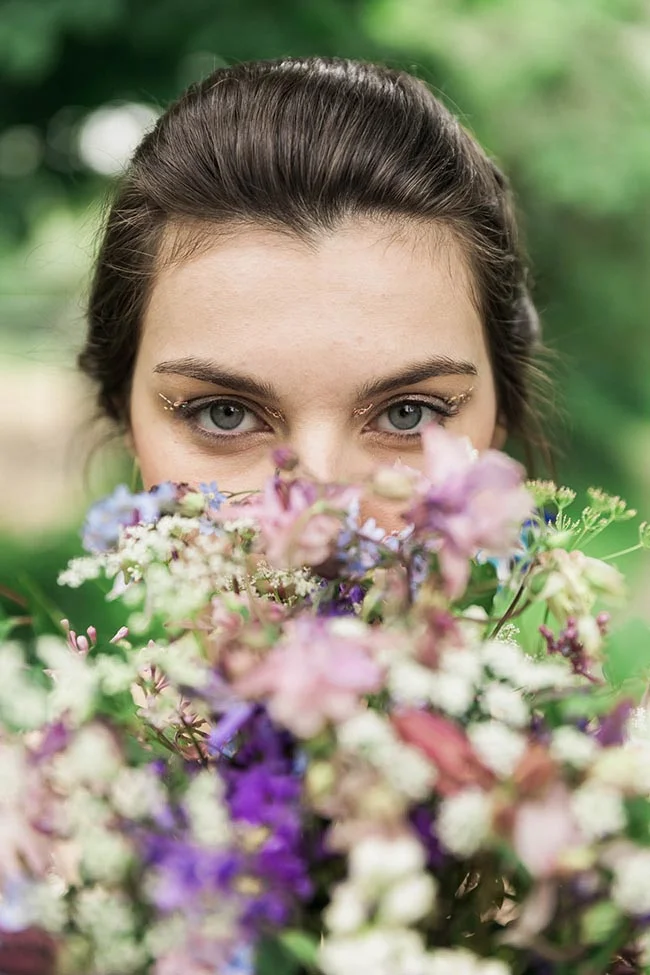 Portrait d'une femme aux yeux clairs cachée derrière un bouquet de fleurs sauvages pastel dans un décor naturel flou.