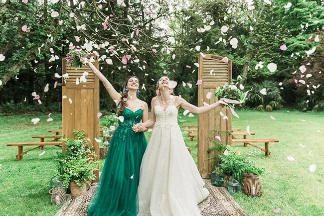 Deux mariées riant sous une pluie de pétales dans un décor forestier avec une robe blanche et une robe vert émeraude.