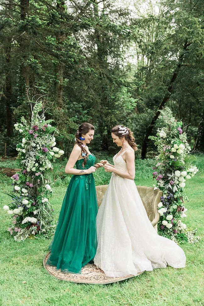 Deux mariées se tiennent la main dans une forêt, l'une en robe verte et l'autre en blanc, entourées de fleurs.