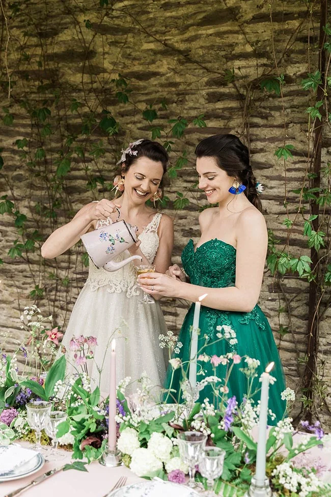 Deux femmes en robes blanche et verte rient près d'une table décorée, devant un mur de pierre et de plantes grimpantes.