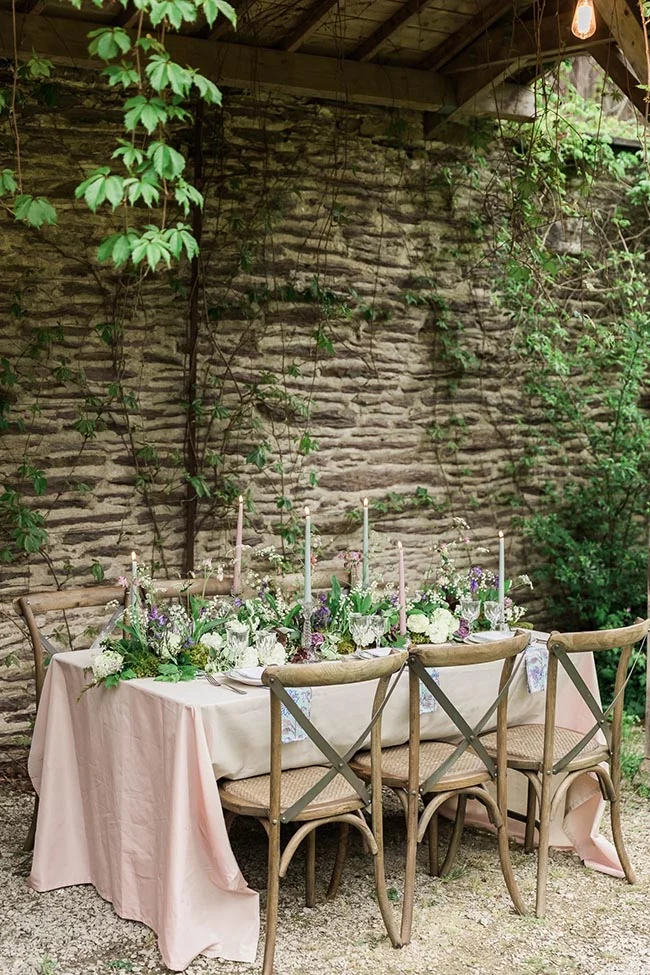 Table élégante nappée de rose avec centre de table floral et bougies, devant un mur de pierre et une végétation luxuriante.