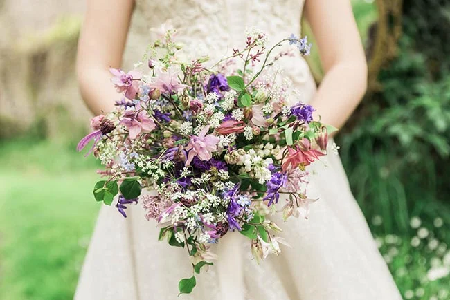 Une mariée tenant un bouquet champêtre aux tons mauve, rose et blanc devant une robe ivoire dans un cadre verdoyant.