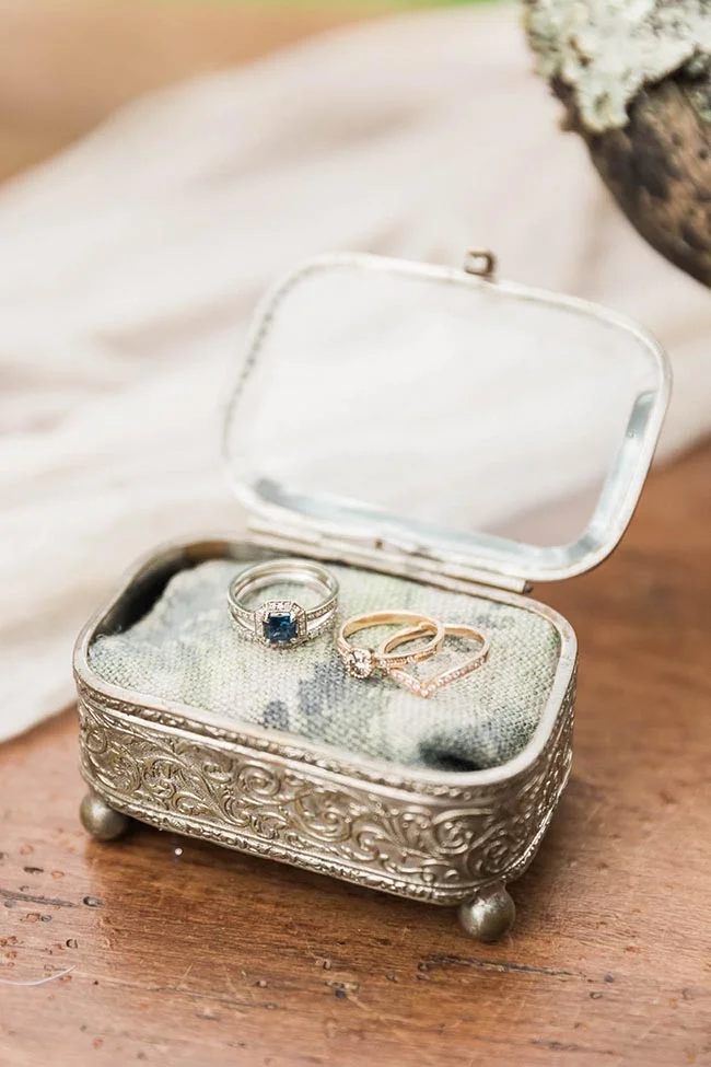 Trois alliances et une bague saphir dans un écrin vintage argenté, posés sur un tissu doux et un bois rustique.