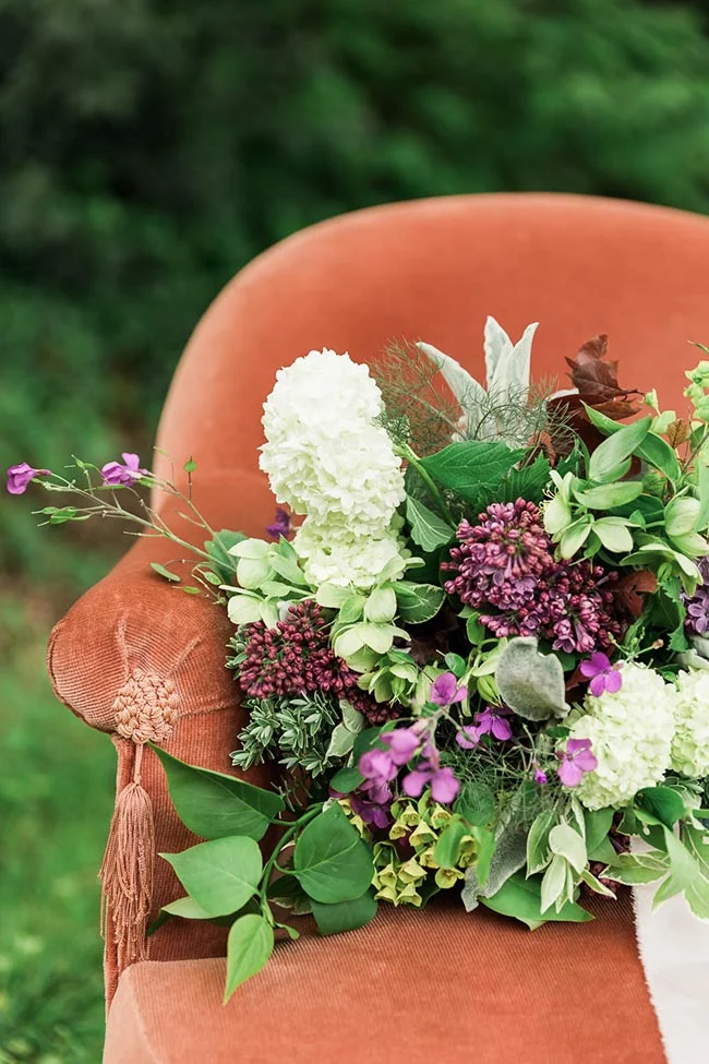 Composition florale aux tons blanc, mauve et vert posée sur un fauteuil en velours terracotta en extérieur.