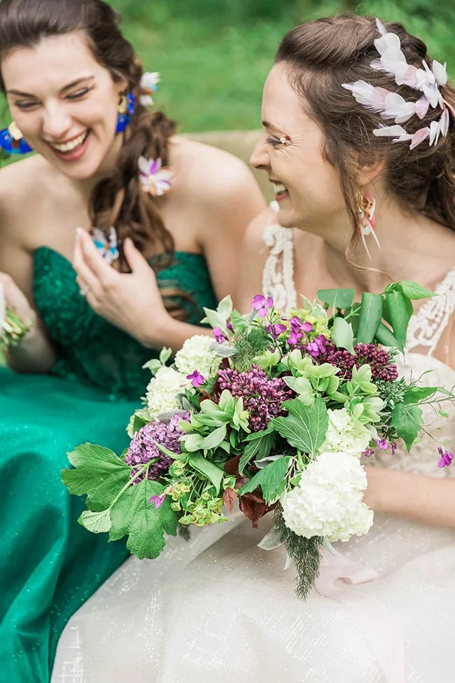 Deux femmes riant ensemble, l'une en robe verte et l'autre en robe blanche avec un bouquet champêtre coloré.