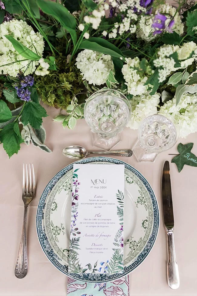 Art de table romantique avec menu, vaisselle bleue et blanche, entourés de fleurs blanches et vertes printanières.