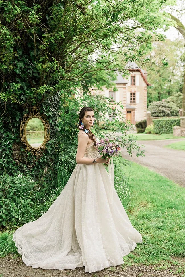 Mariée en robe crème avec bouquet champêtre, posant dans un jardin luxuriant devant un château et un miroir doré.