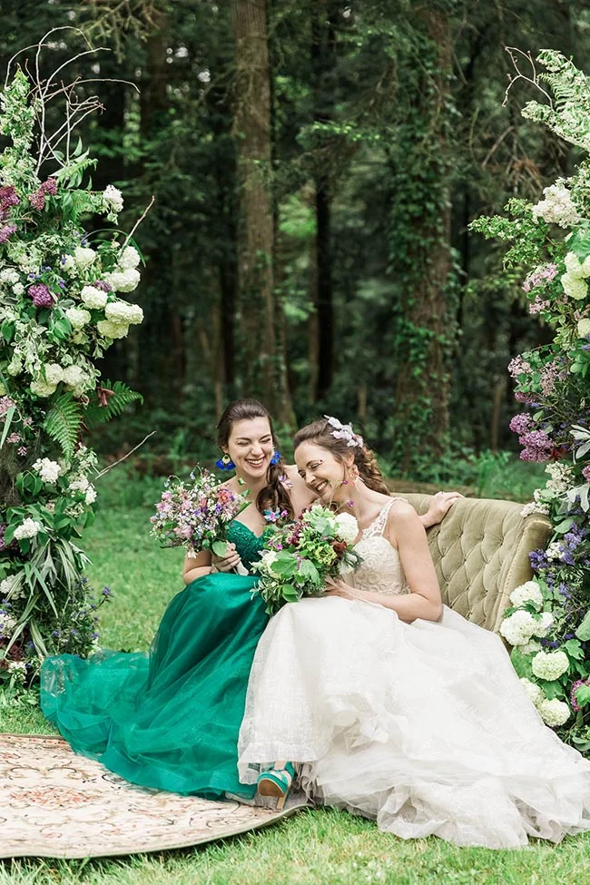 Deux mariées riant dans une forêt, l'une en robe blanche et l'autre en robe verte, entourées de fleurs et de verdure.