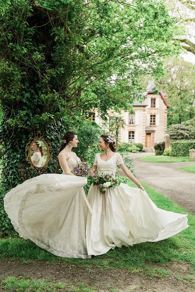 Deux mariées en robes blanches et bouquets colorés s'amusent dans un jardin luxuriant devant un château.