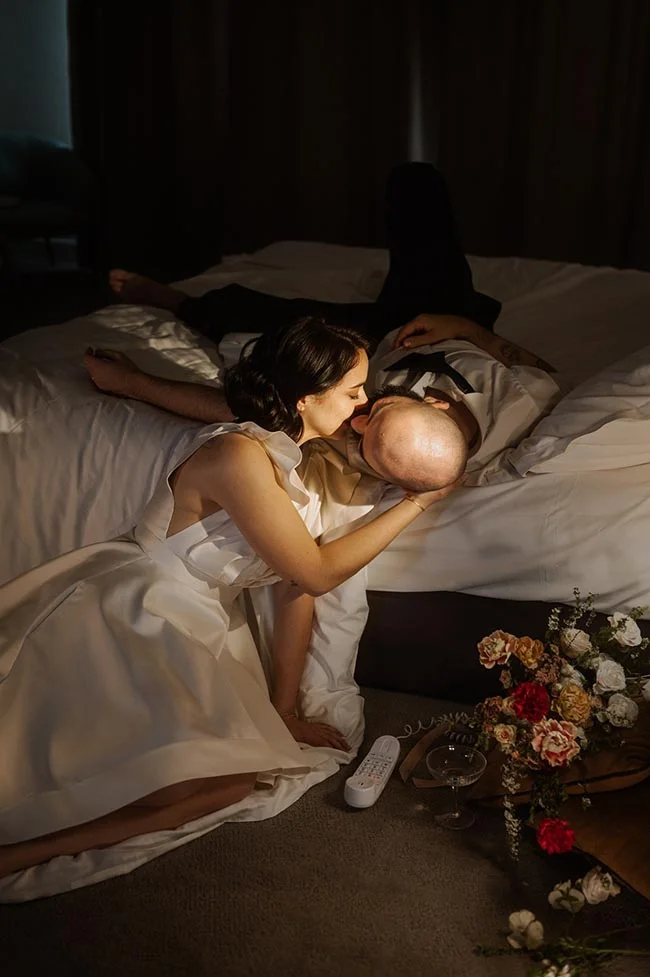 Couple en tenue de mariage s'embrassant tendrement sur un lit dans une ambiance tamisée et intimiste.