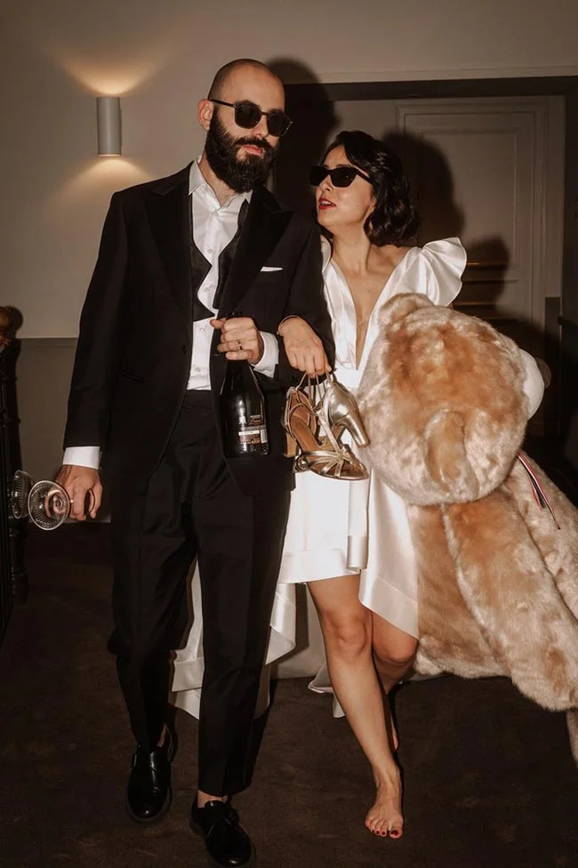Couple chic en tenue de mariage et lunettes noires, avec champagne et manteau en fourrure, ambiance festive et décalée.