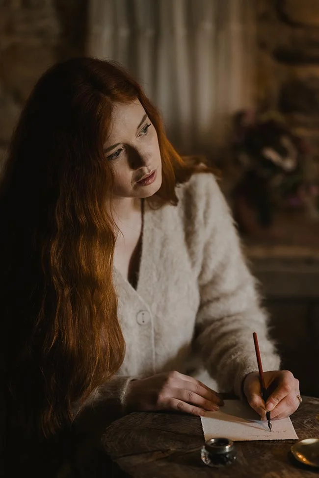 Jeune femme aux cheveux roux écrivant une lettre à la lumière d'une bougie dans une ambiance intime et romantique.