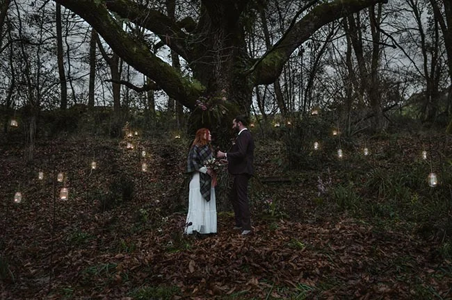 Couple en tenue de mariage sous un grand arbre dans une forêt sombre, entouré de lanternes suspendues et de feuilles mortes.