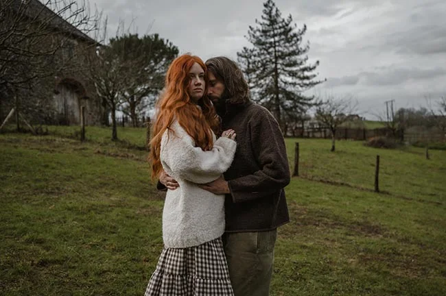 Couple enlacé dans un pré verdoyant sous un ciel gris, la femme aux longs cheveux roux portant un pull blanc.