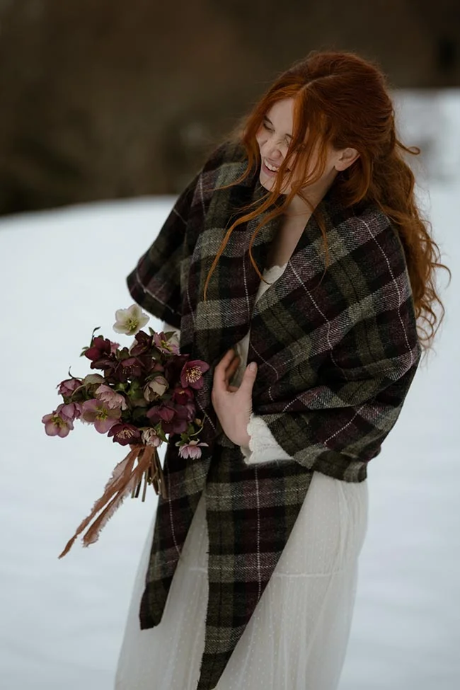 Mariée rousse souriante en robe blanche et plaid à carreaux sombres, tenant un bouquet hivernal dans la neige.