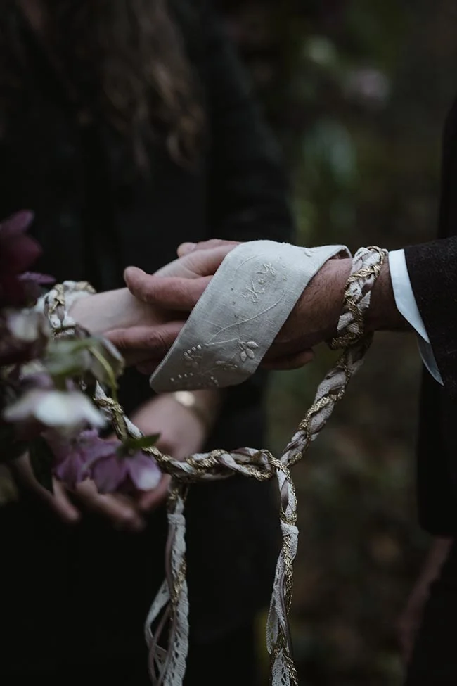 Gros plan sur deux mains liées par un ruban tressé blanc et or, dans une ambiance romantique et sombre.