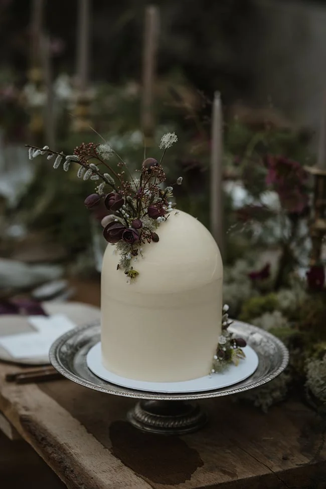 Gâteau de mariage minimaliste blanc crème, décoré de fleurs sauvages sombres, sur un plateau argenté rustique.