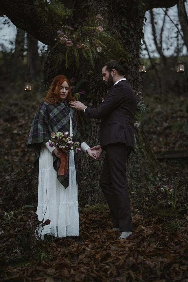 Couple en tenue élégante et châle écossais se tenant la main dans une forêt sombre et romantique.