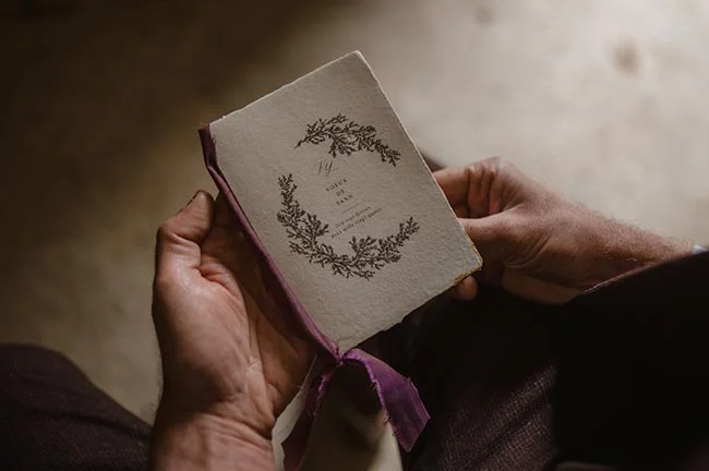 Gros plan sur des mains tenant un petit carnet blanc à couverture illustrée et ruban violet.