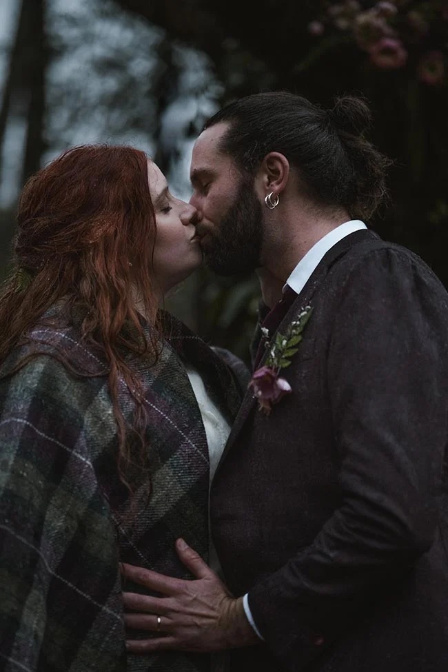 Un couple s'embrasse tendrement en extérieur, elle en cape à carreaux et lui en costume sombre, dans une ambiance feutrée.