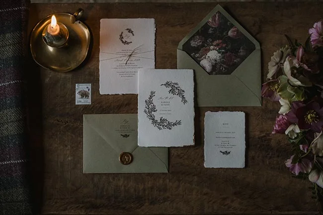 Papeterie de mariage élégante aux tons neutres et motifs floraux, posée sur du bois sombre avec une bougie allumée.