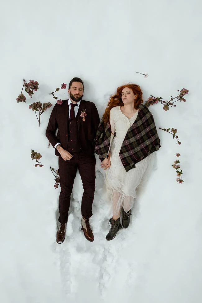 Couple marié allongé dans la neige, entouré de fleurs, dans une ambiance romantique et hivernale.