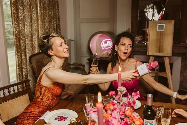 Deux femmes en robes colorées s'amusent lors d'un EVJF festif autour d'une table décorée de fleurs et de champagne.