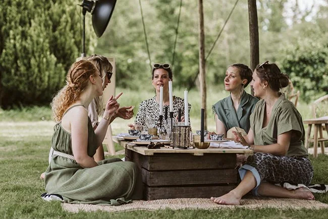 Quatre femmes discutent joyeusement autour d'une table basse en bois lors d'un pique-nique chic en plein air.