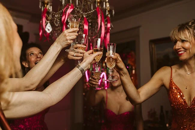 Des amies en robes pailletées trinquent avec des coupes de champagne sous un lustre décoré de rubans roses.