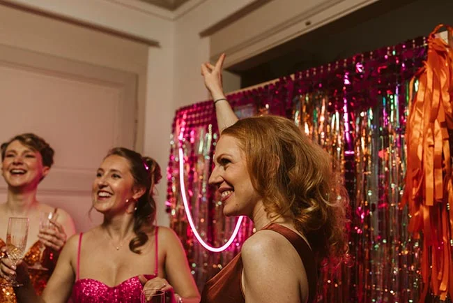 Trois femmes souriantes font la fête devant un rideau à paillettes roses et violettes dans une ambiance festive.