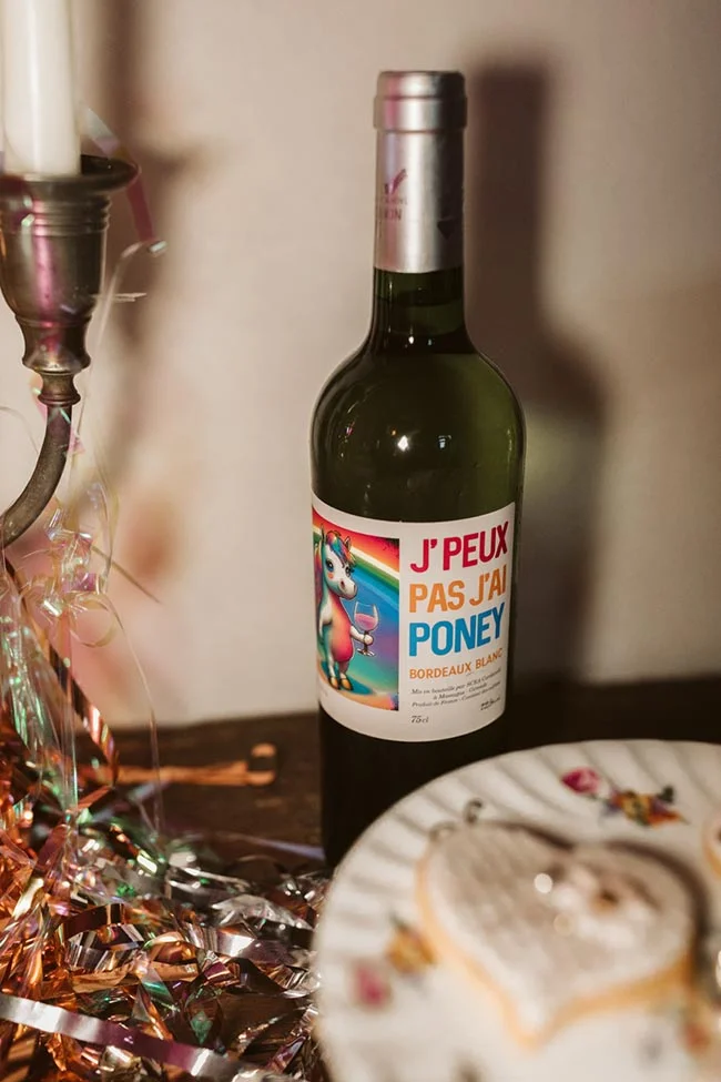 Bouteille de vin blanc humoristique entourée de confettis brillants et d'un gâteau en forme de cœur.