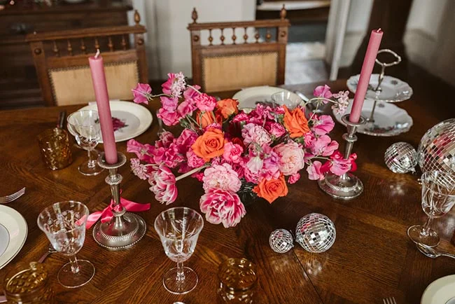 Table en bois dressée avec un bouquet de fleurs roses et orangées, des bougies roses et des verres en cristal.