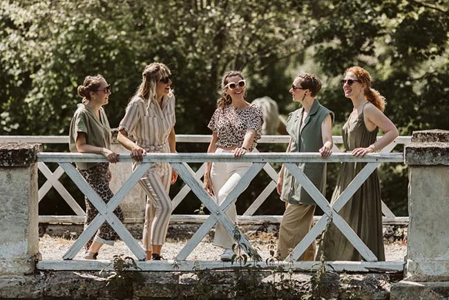 Cinq amies souriantes et élégantes posent sur un pont blanc dans un cadre verdoyant et lumineux.