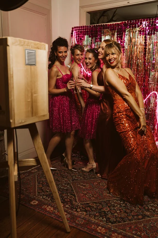 Cinq femmes souriantes en robes festives roses et orange posent devant un rideau à paillettes près d'un photobooth.