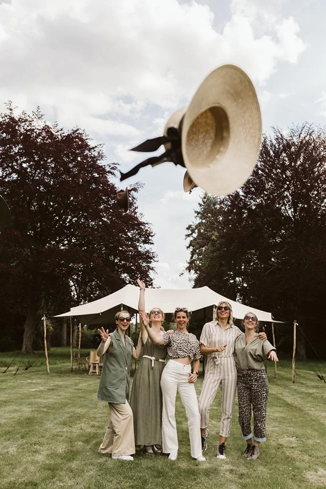 Cinq amies souriantes en tenues beiges et vertes devant une tente blanche, un chapeau de paille volant dans les airs.