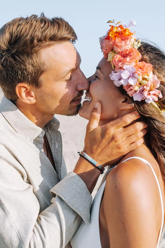 Couple amoureux s'embrassant tendrement sur une plage, la mariée portant une couronne de fleurs colorées.