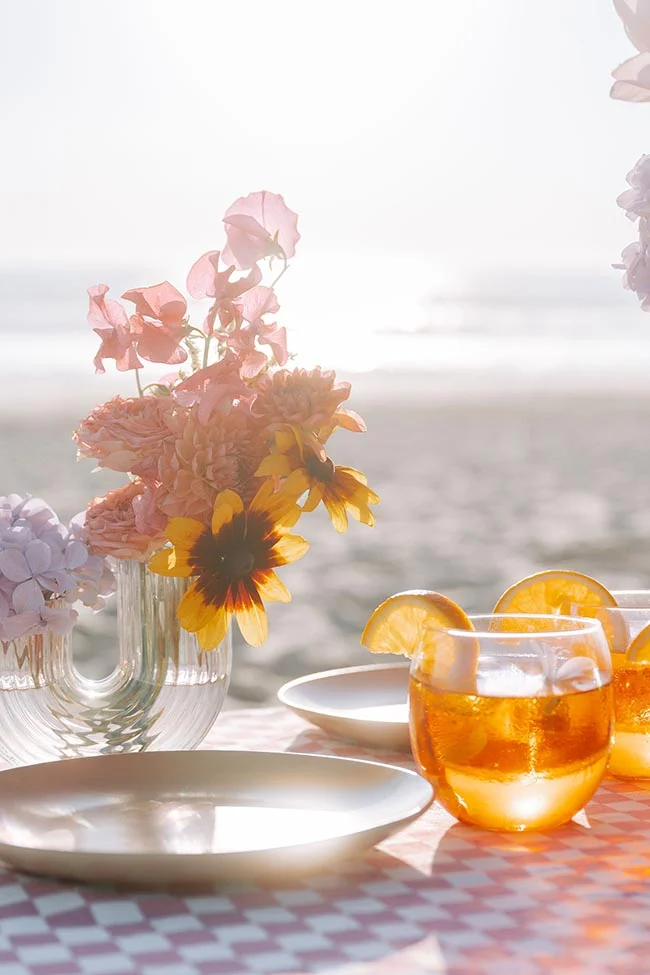 Composition lumineuse de fleurs roses et jaunes et cocktails à l'orange sur une table à carreaux face à la plage.
