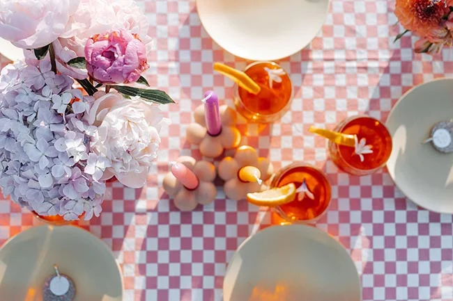 Vue aérienne d'une table de fête avec nappe à carreaux roses, fleurs blanches et roses, et cocktails colorés.