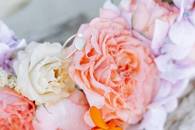 Bagues de mariage posées sur un bouquet de roses pastel et fleurs mauves dans une ambiance douce et romantique.