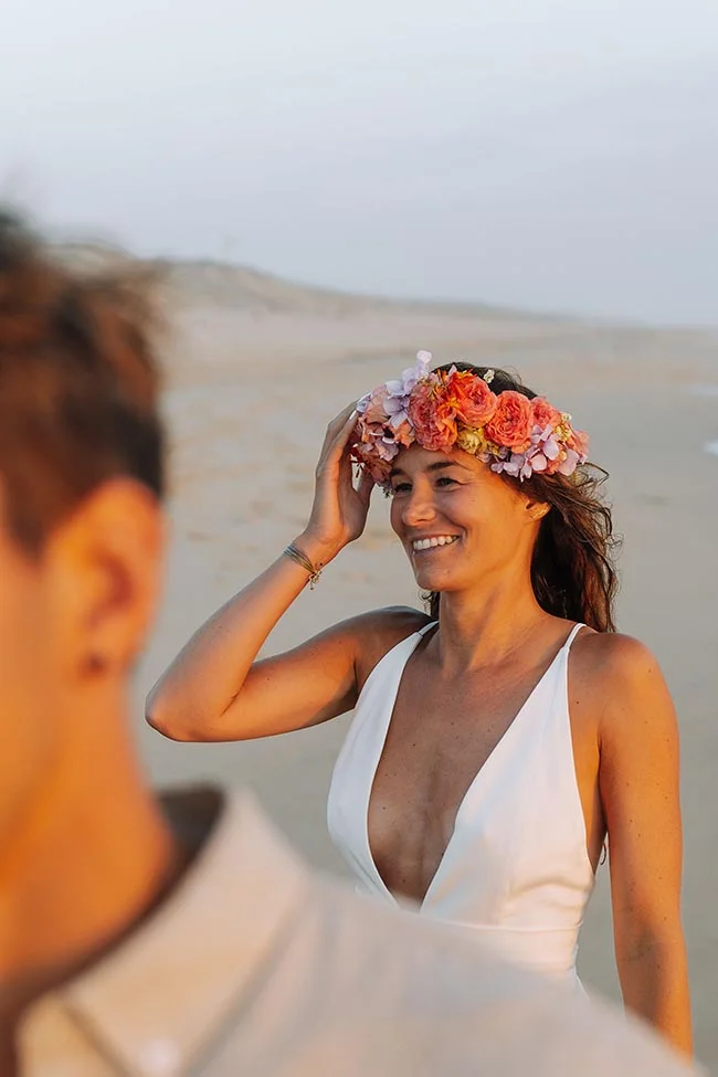 Mariée souriante avec une couronne de fleurs colorées, portant une robe blanche sur une plage au coucher du soleil.