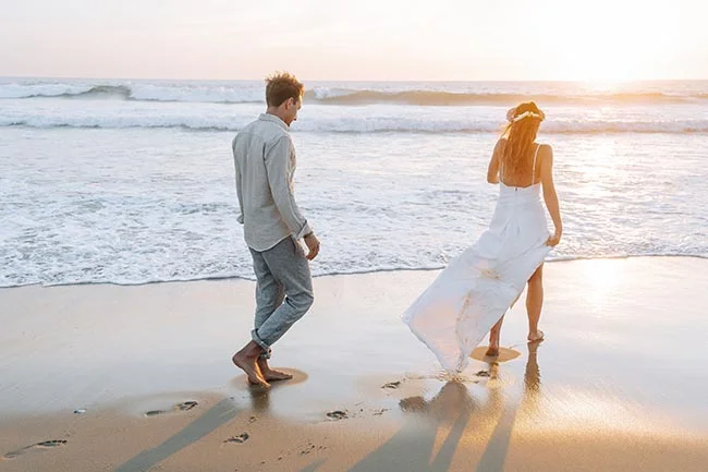 Couple en tenue blanche marchant pieds nus sur une plage au coucher du soleil, ambiance romantique et lumineuse.