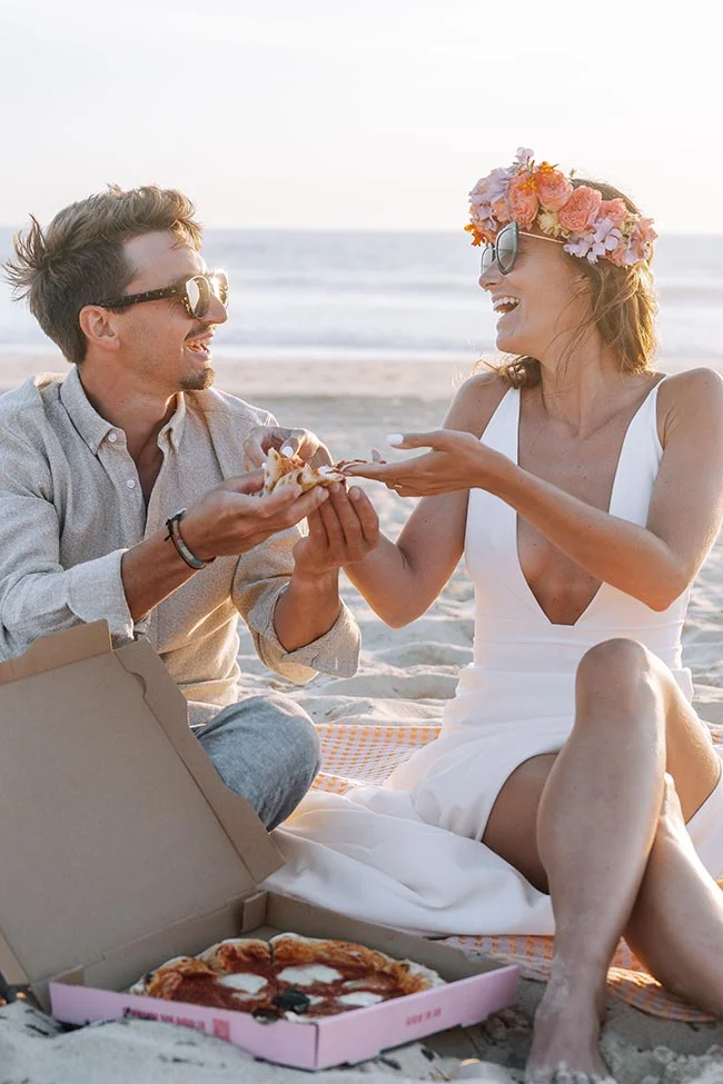 Jeunes mariés souriants partageant une pizza sur le sable lors d'un cocktail décontracté à la plage au coucher du soleil.