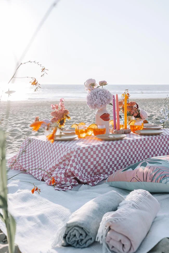 Pique-nique chic sur la plage avec nappe à carreaux roses, fleurs colorées et cocktails au coucher du soleil.