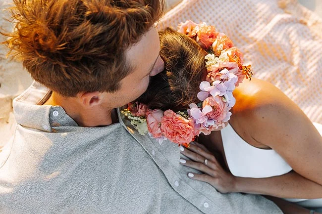 Couple s'enlaçant tendrement sur le sable, la mariée portant une couronne de fleurs colorées dans une ambiance solaire.