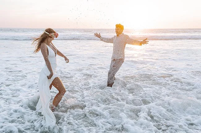 Jeunes mariés joyeux s'amusant dans l'écume d'une plage au coucher du soleil, ambiance romantique et lumineuse.
