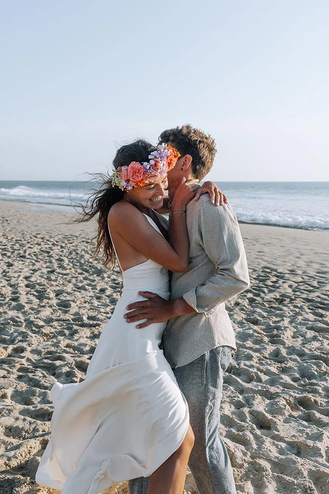 Couple marié s'enlaçant avec tendresse sur une plage de sable blanc, la mariée portant une couronne de fleurs colorées.